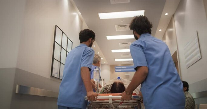 Backview Tracking Shot of Group of Medical Specialists Transporting an Elderly Patient with a Stretcher Through a Hospital Corridor. Health Clinic Receiving an Emergency Case for Intensive Care Unit