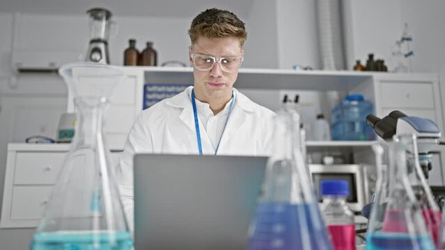 Stressed young caucasian male scientist in doubt, taking off glasses while working on laptop, thinking hard over problem in laboratory.