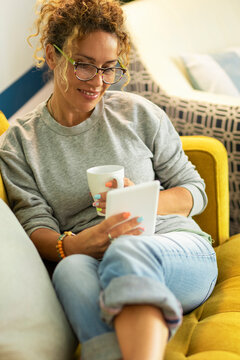 Pretty woman enjoy relax at home reading a book tablet reader and drinking a tea or coffe sitting on a yellow sofa in living room - modern online and female people using device gadget to buy and read