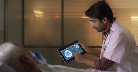 Hospital Ward: Young Indian Male Doctor Talks to Elderly Patient Resting in Bed, Uses Tablet Computer, Shows and Explains Brain CT MRI Scan Results. Physician Talks to a Man Recovering after Surgery