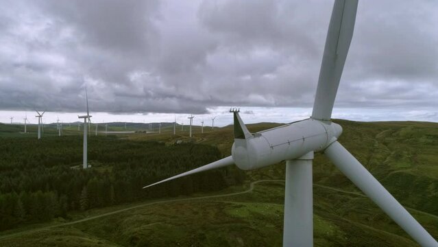 Aerial drone slow track shot of wind turbine with reveal of whole wind farm, cloudy day, forest surroundings. 
