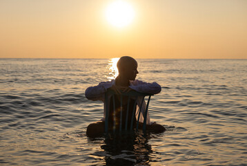 beautiful woman with alopecia relaxing on seashore,sitting or dragging chair in water,sunset or...