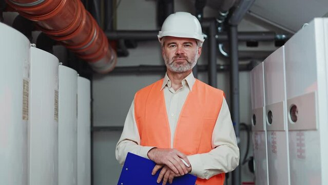 Gray haired male construction worker making on clipboard while standing in his indoor workspace. Skillful mature professional standing next to machines and feeling responsibility during day shift. - Powered by Adobe