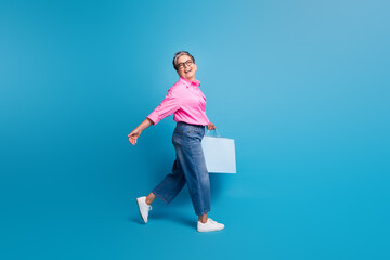 Full size photo of carefree positive person with grey hair go shopping in empty space hold new clothes isolated on blue color background