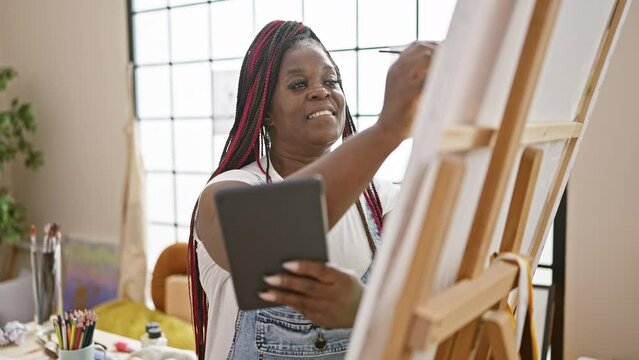 Confident, beautiful african american woman artist happily drawing on touchpad, bringing creativity to life in modern art studio setting.