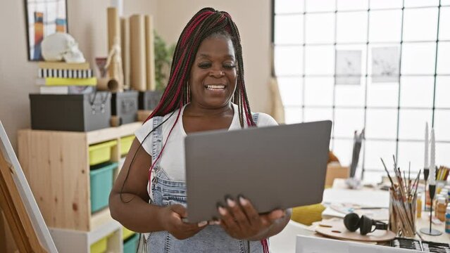 Confident african american woman artist with braids, smiling as she paints on canvas and uses laptop in a cozy art studio