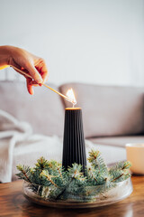 Female hand lighting a candle with a match in winter cozy composition for hygge home mood. Black candle and spruce branches on the coffee table in living room. Natural Christmas decorations for home.