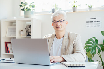 Portrait of smiling grey hair middle-aged man working on laptop while sitting at his work place in...