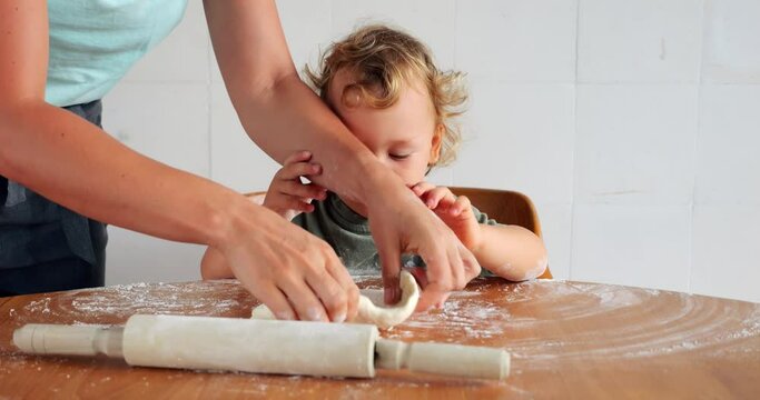 Mom and baby boy share messy, homemade pizza preparation moment in kitchen. As mom handles dough, toddler eagerly helps, even playfully trying to take charge. Heartwarming scene at home