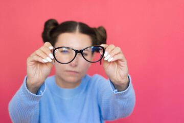 a young pretty woman in a blue sweater with poor eyesight holds glasses in her hands, squints and looks through them into the camera. Vision problems. Ophthalmology - obrazy, fototapety, plakaty