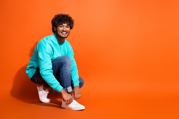 Full length photo of cheerful indian guy advertising new sneakers adidas brand lacing up sitting floor isolated on orange color background