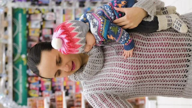 Vertical video. Young woman with cute baby shopping for Christmas decorations at a store. Winter holidays concept