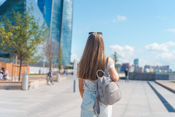 A beautiful young girl in jeans with long hair with a backpack walks down the street with a view of...