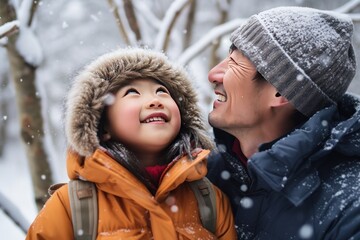 Fototapeta premium Asian father and daughter warmly dressed in winter during snowfall embrace with smiles. Asian dad warmly hugs little daughter in nature during snow. Asian family warm in cold with loving hugs