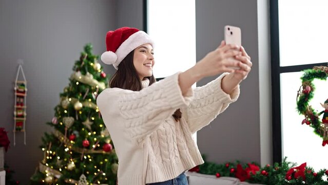 Young beautiful hispanic woman make selfie by smartphone standing by christmas tree at home