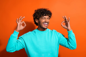 Portrait of handsome funky handsome man eye wink blink fingers demonstrate okey symbol isolated on orange color background