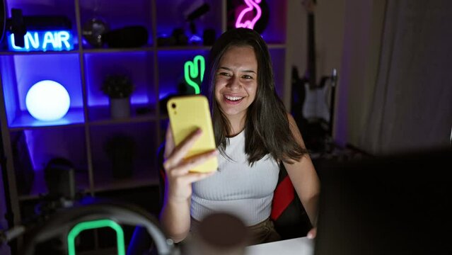 Smiling young hispanic woman, streaming a thrilling video game, engrossed in digital entertainment from her neon-lit gaming room during a lively video call