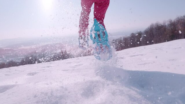 LENS FLARE, SLOW MOTION, CLOSE UP: Playful young woman is running with snowshoes in freshly fallen snow on a beautiful sunny day. Following a lady as she runs downhill in snowshoes and sprays snow.