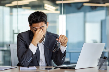 Overtired and overwhelmed businessman at workplace inside office, man took off glasses rubbing...