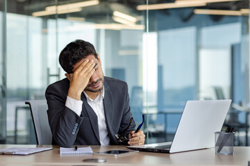 Overtired and overwhelmed businessman at workplace inside office, man took off glasses rubbing eyes, dizziness migraine and headache, man in business suit working late with laptop at workplace. - obrazy, fototapety, plakaty