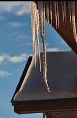 Vertical closeup of ice dams hanging from the edges of a roof against a blue sky, winter background