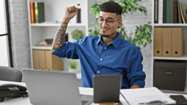 Young, tattooed latin man ecstatically celebrates business success at the office, crossing fingers, working on laptop and touchpad