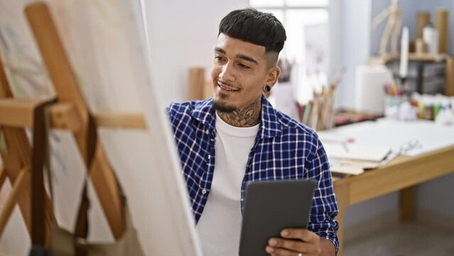 Confident young latin man, a tattooed artist, drawing on touchpad while smiling and sitting at his art studio class indoors