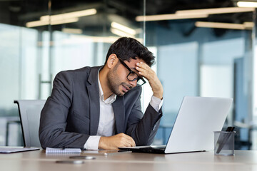 Depression at workplace, businessman disappointed with achievement results sad in despair inside office, man in business suit unhappy working with laptop. - obrazy, fototapety, plakaty