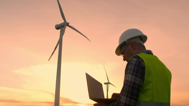 Man maintenance engineer works at wind farm at sunset with a laptop in his hands