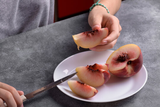 Authentic female hands holding a piece of a fresh ripe peach on white plate on a grey table background, close up. Healthy summer snack, food lifestyle - Powered by Adobe