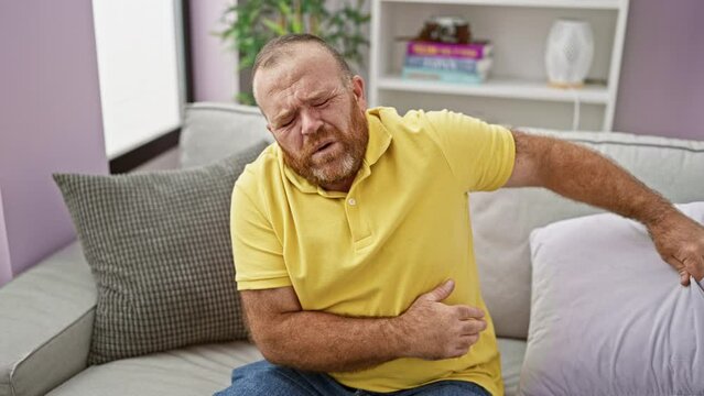 Worried, handsome, middle-aged caucasian man suffering a serious stomach ache, lying unhappily on the sofa at home. indoor portrait of ill male with beard, forced to rest due to the distressing pain.