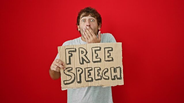Young man holding cardboard free speech banner covering mouth with hand, shocked and afraid for mistake. surprised expression over isolated red background