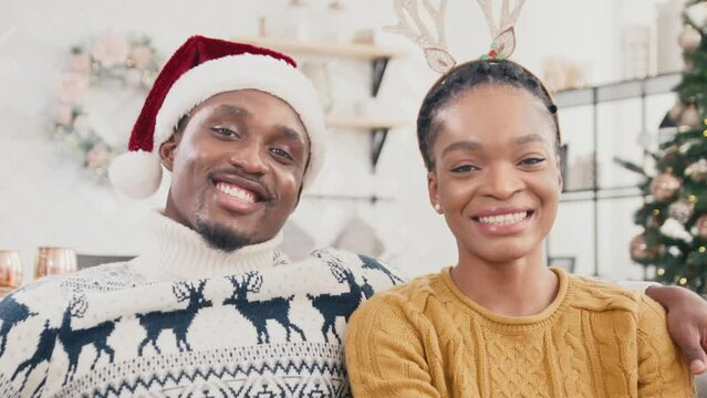 Portrait of African American married couple wearing knitted sweaters sitting on sofa. Attractive man with Santa Claus hat hugging his pretty wife which in headband of deer horns. Christmas spirit.