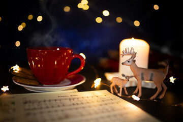 Warm toned Christmas still life with red coffee cup, carol music sheet and deer figurine in candle...