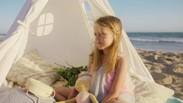 Little happy girl telling interesting story holding in hands stuffed doll toy. Adorable child funny face at ocean beach in golden sunset light. Cute little girl with long blond hair flying at wind 4K