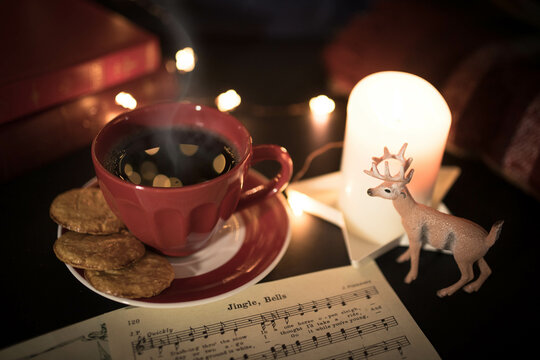 Warm toned Christmas still life with red coffee cup, carol music sheet and deer figurine in candle lit setting. High angle view.