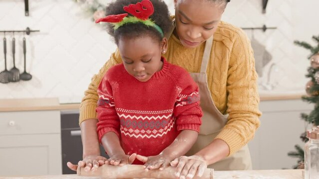Happy African American little daughter wearing headband of deer horns while helping her adorable mother rolling out dough in modern kitchen. Caring mommy teaching her child cooking Christmas cookies.