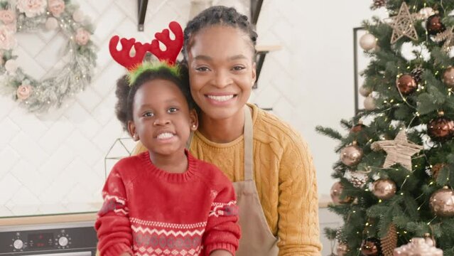 Pretty African American mother with her daughter standing in kitchen next to Christmas tree. Happy woman with apron. Hugging her lovely child in headband of deer horns. Preparing Christmas foods.
