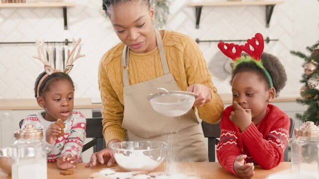 Beautiful African American mother sprinkling powdered sugar on gingerbread cookies while her little children eating tasty baking in kitchen. Happy family spending time together during cooking food.