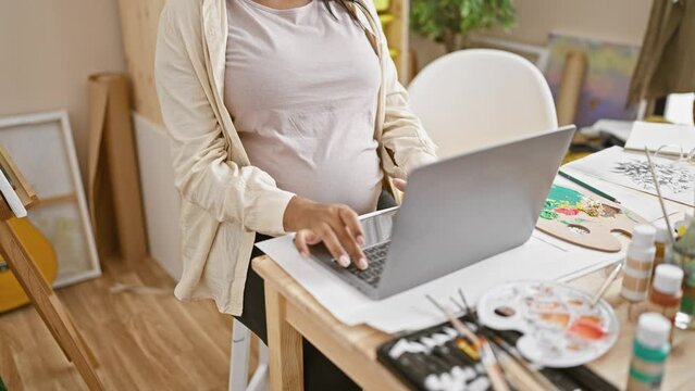 Eager young pregnant woman sitting at her art studio table mastering her craft, painting on canvas with laptop as guide.