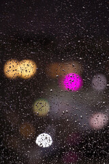 Rain drops on window glass at night with blurred bokeh light close up. Unfocused colorful abstract lights on black background texture