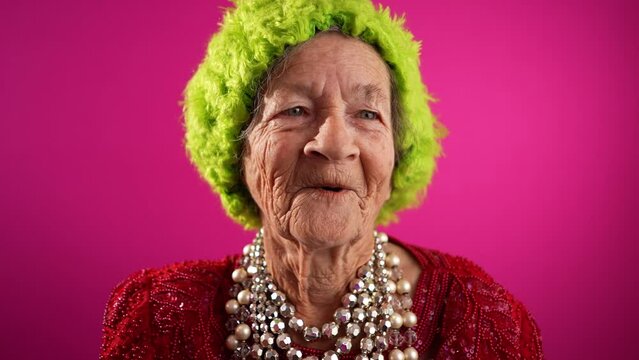 Smiling fisheye portrait caricature of funny elderly woman smiling with green wig or hat and no teeth isolated on pink background. Slow motion