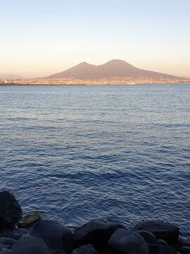 The calm blue waters surrounding Naples are bordered by rocks in the foreground and Mt Vesuvius in the distance. The sky is clear. A few boats are on the water.
