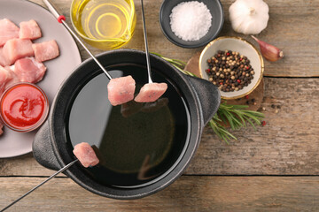 Fondue pot with oil, forks, raw meat pieces and other products on wooden table, flat lay