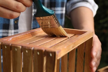 Man applying varnish onto wooden crate against blurred background, closeup