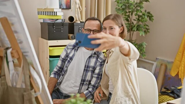 Two smiling artists immersed in taking a selfie with smartphone amidst the colorful chaos of their art studio