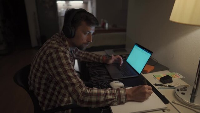 Focused man in headphones with a laptop late at night at home, taking notes, student learning a language, watching an online webinar, hearing an audiocourse, the concept of e-education. Homework. 