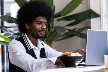 Young afro boy sitting at the bar of a restaurant while working on his laptop.