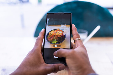 Two black hands of a man holding a mobile phone while he is taking a picture to his healthy food. Poke bowl.