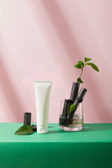 Glass cup containing bamboo charcoal and green tea leaves. A white cosmetic tube is placed on a green table with a pink background. Concept of natural cosmetics.